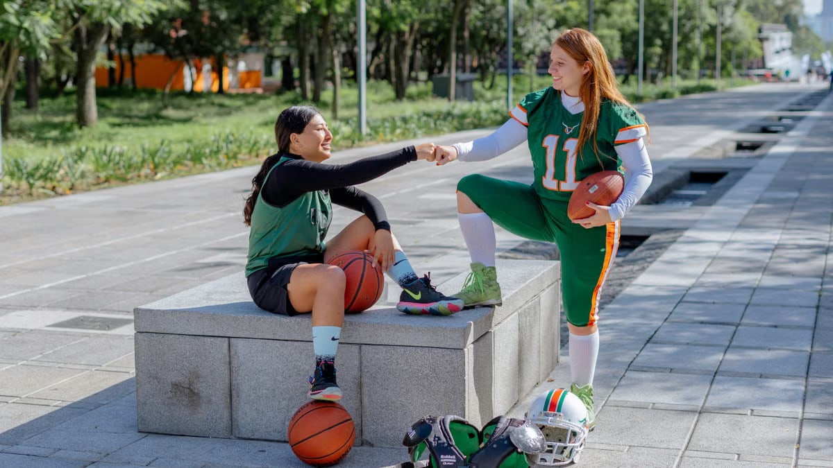 Two female athletes share a fist bump while sitting outdoors, holding sports equipment in Mexico City.